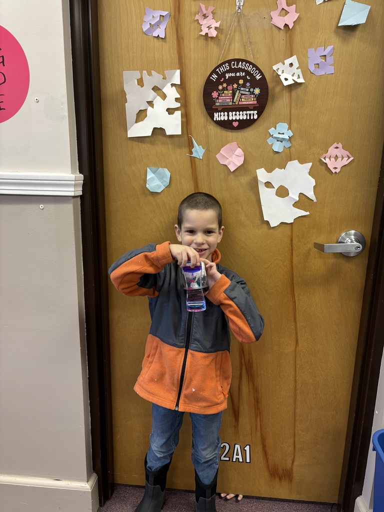 A student stands in front of a classroom door holding up a finished winter craft jar, smiling while wearing a jacket and boots.