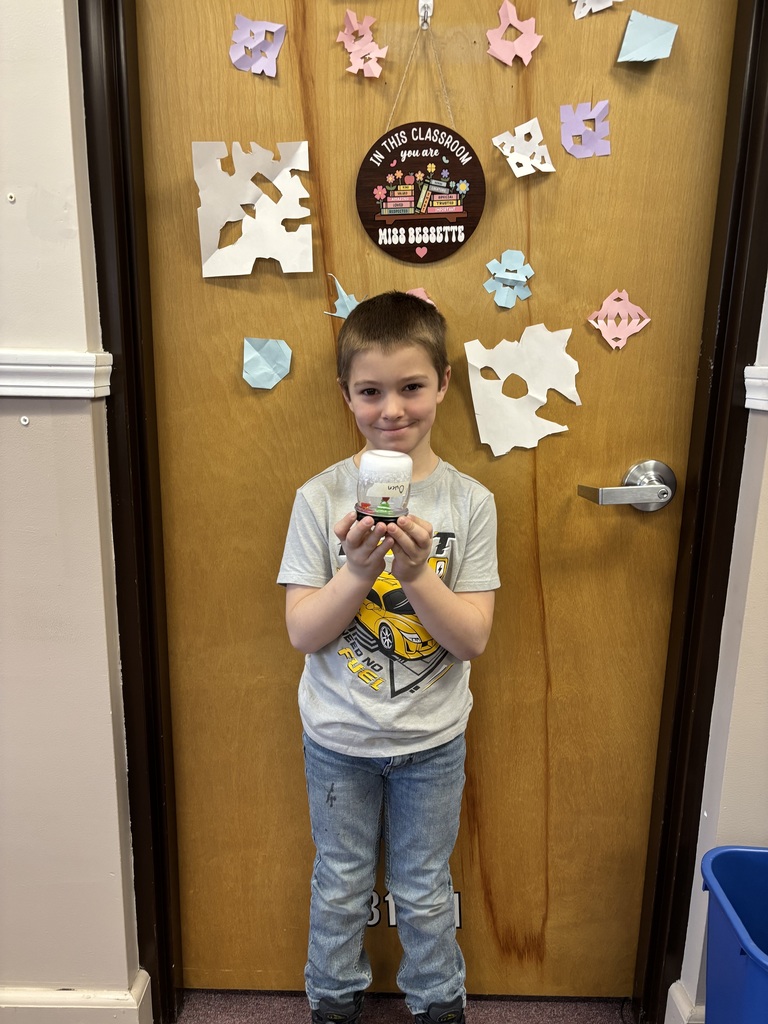 A student stands in front of a classroom door decorated with paper snowflakes, holding a completed winter craft jar and smiling.