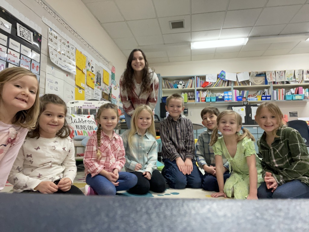  In the image, a group of eight elementary school children and their teacher are posed together in a classroom. The teacher, a woman with long brown hair, stands in the center back of the group, wearing a red and white plaid shirt.  The children, four girls and four boys, are sitting or kneeling on the floor in front of her. They are all smiling at the camera