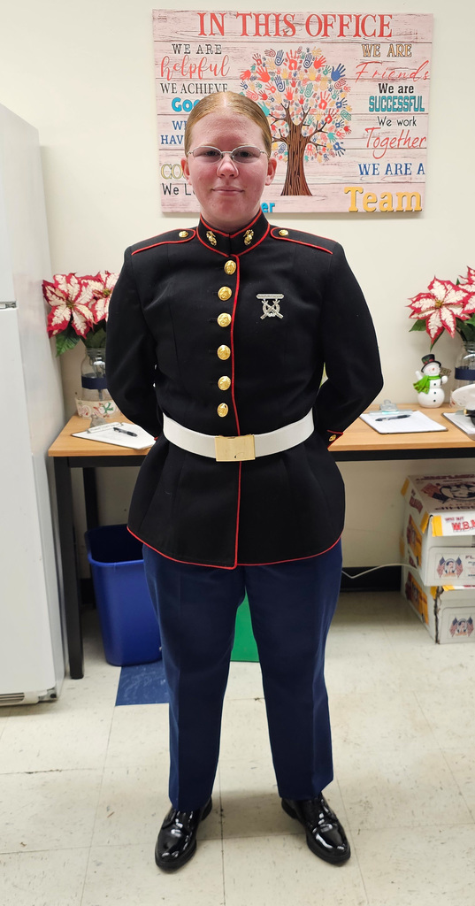 A Slate Valley graduate stands indoors wearing a formal military dress uniform with dark jacket, brass buttons, white belt, and navy trousers. She is standing with hands behind her back in a school office area with a refrigerator, tables, poinsettias, and a wall sign in the background.