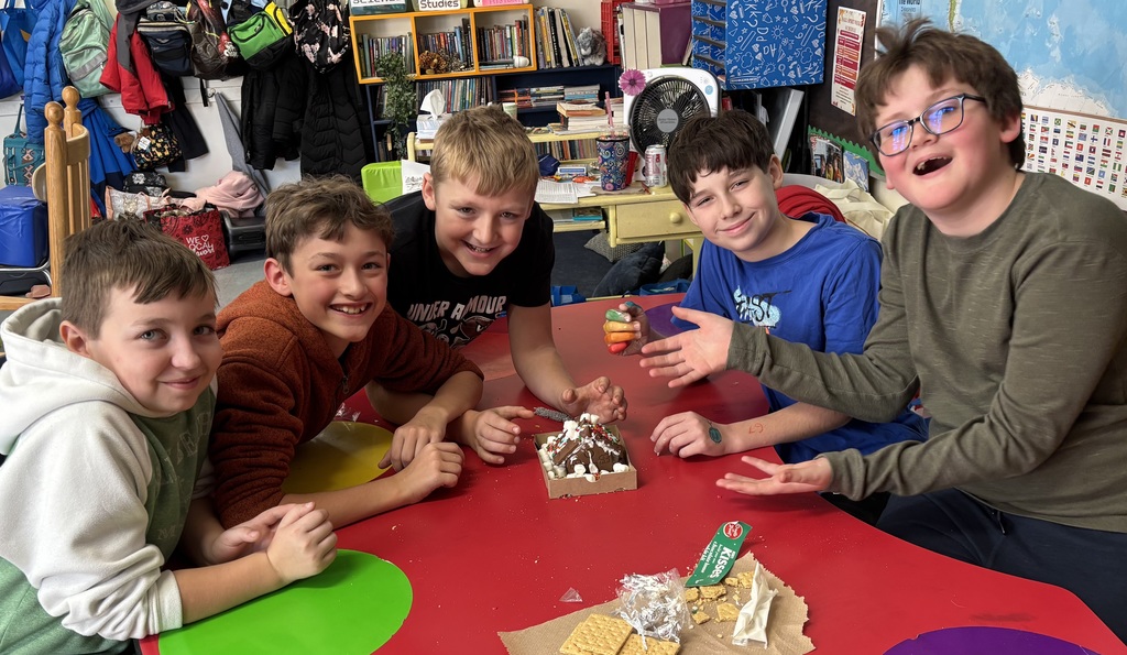 Five students pose proudly around a completed gingerbread house on a red table, gesturing toward their decorated creation.