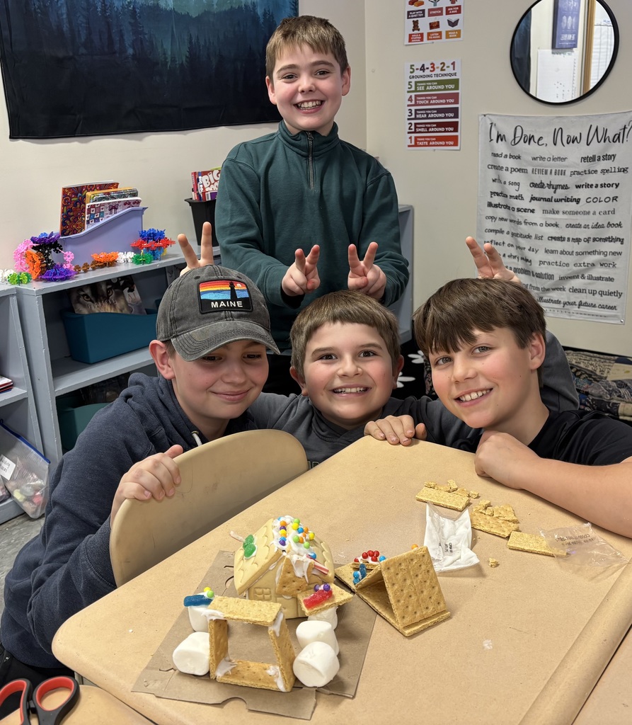 Four students grin behind a gingerbread house with marshmallow supports, showing playful hand gestures in a classroom.