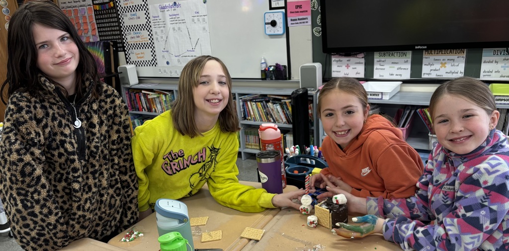 Four students smile behind a gingerbread house with candy decorations while sitting together at a classroom table.