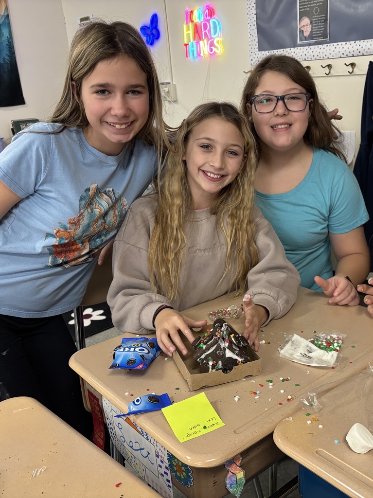 Three fifth-grade students smile at a desk while decorating a gingerbread house covered in icing and colorful candy.