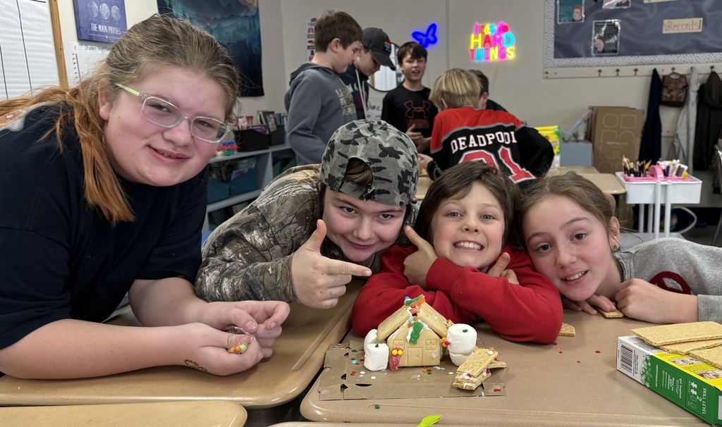 Four students lean close to a gingerbread house on a desk, smiling as they display their finished project.