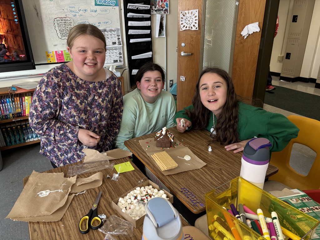 Three students gather around a table displaying a finished gingerbread house, with craft supplies, candy, and icing spread across the desks.