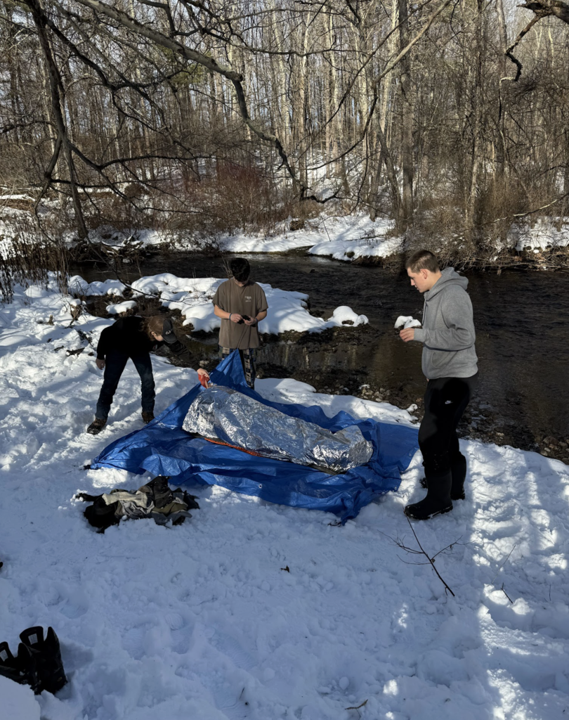 Students spread out a blue tarp on snowy ground beside a creek, working together to set up a shelter base in a wooded area.