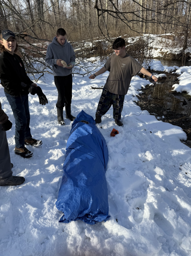 A group of students stands in the snow near a creek while a blue tarp lies on the ground, preparing materials for a winter shelter or rescue simulation.