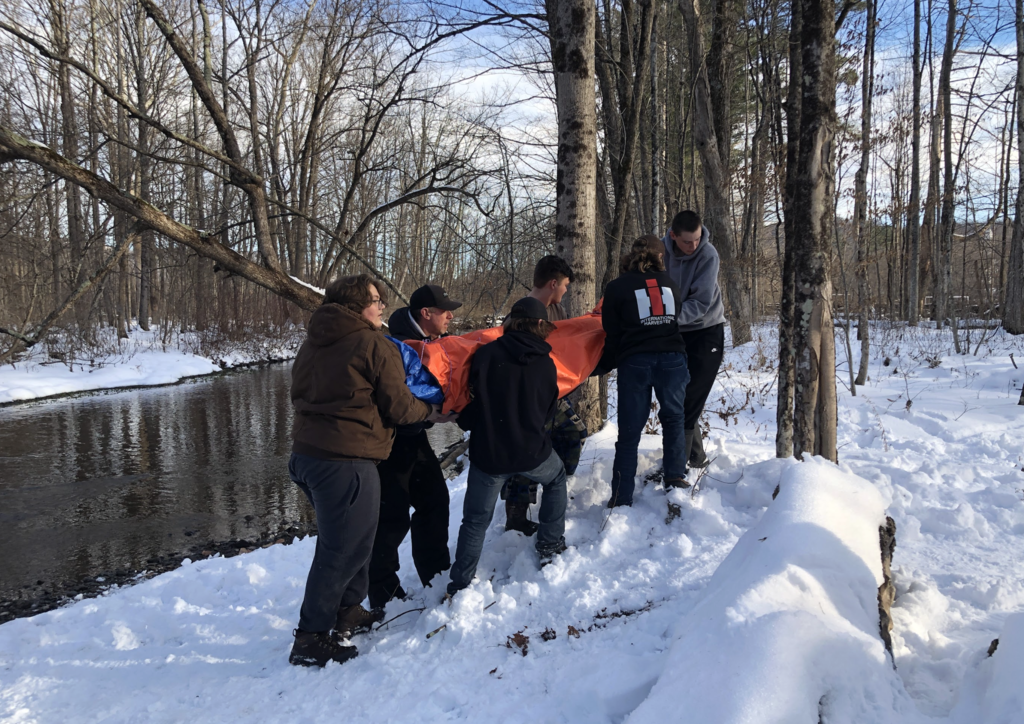A group of students carefully carries a wrapped classmate on a snowy slope near a creek, demonstrating teamwork during a simulated rescue.