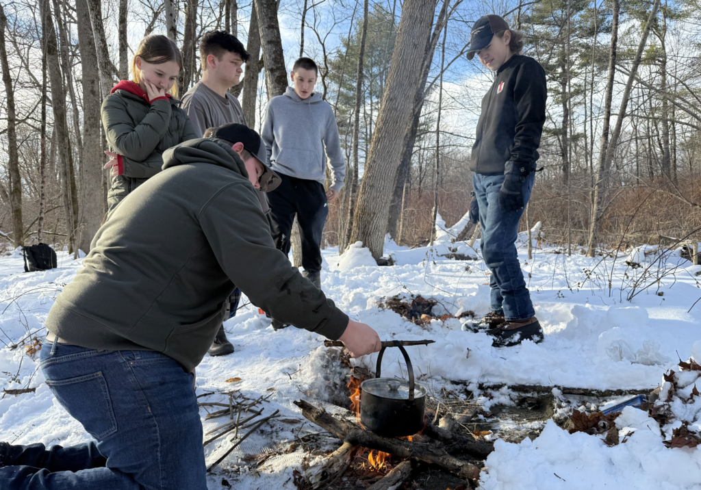 Students gather around a small campfire in the snow as one student stirs a pot hanging over the fire during an outdoor cooking lesson.