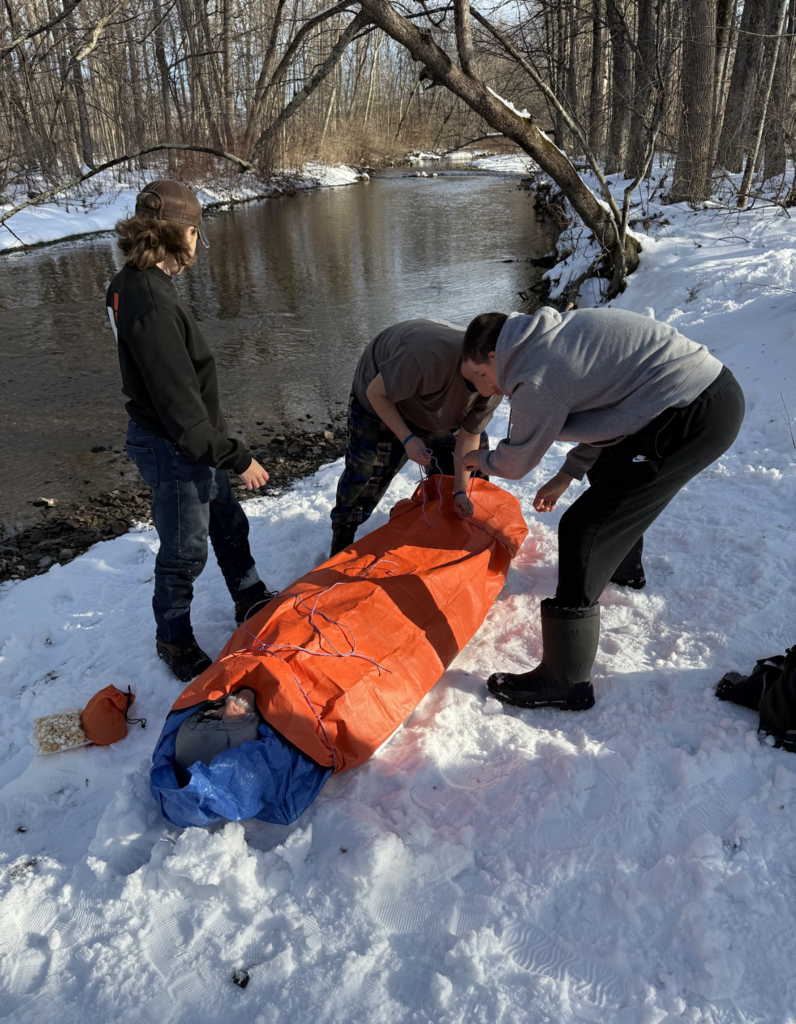 Students wrap a classmate in an orange emergency bivy while standing on snow near a creek, practicing cold-weather safety techniques.