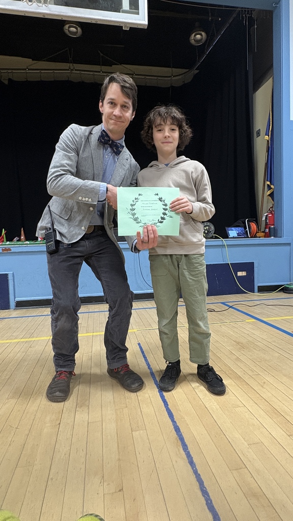 A teacher stands next to an elementary student holding a green PBIS classroom award certificate during a school assembly on the gym stage.