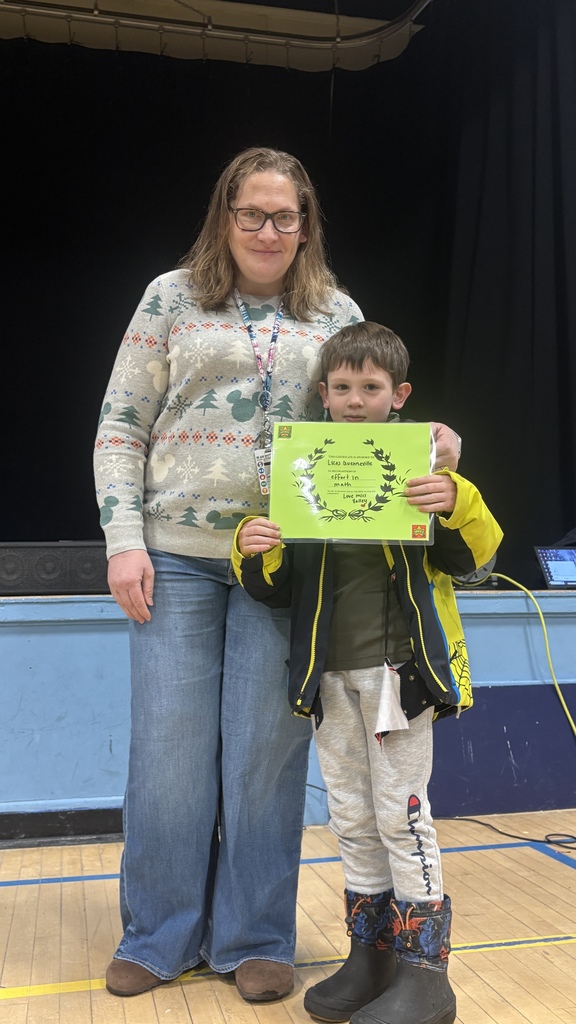 A teacher stands beside an elementary student holding a green PBIS classroom award certificate, both smiling on the school stage.