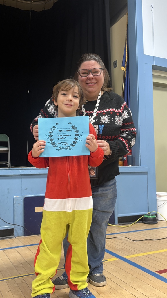 An elementary student in a colorful outfit holds a blue PBIS classroom award certificate while standing in front of a teacher on the school stage.