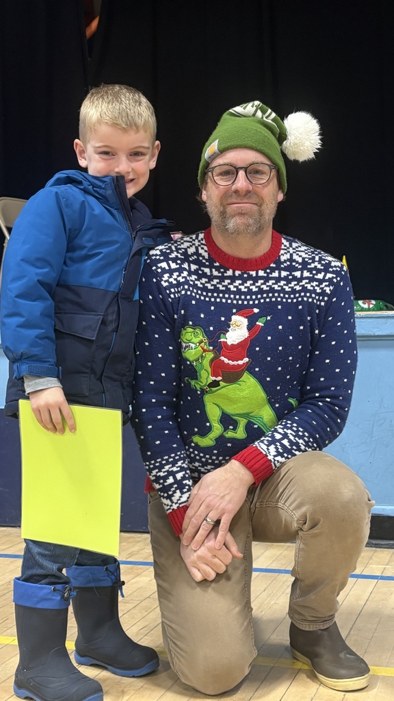 An elementary student stands smiling next to a staff member wearing a festive holiday sweater, holding a bright award certificate during a PBIS assembly on the school stage.