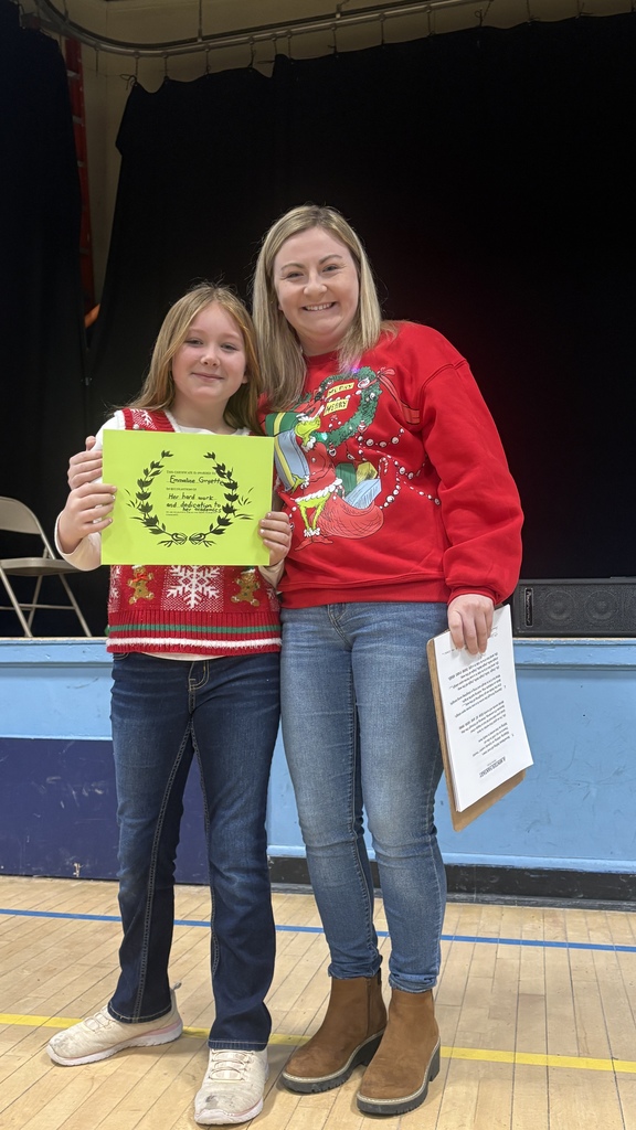 An elementary student holds a green PBIS classroom award certificate while standing next to a teacher wearing a red holiday sweater during a school assembly.