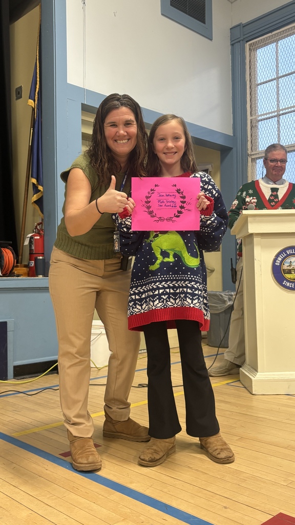 A teacher gives a thumbs-up while standing beside an elementary student holding a pink PBIS classroom award certificate, with an administrator visible at a podium in the background.