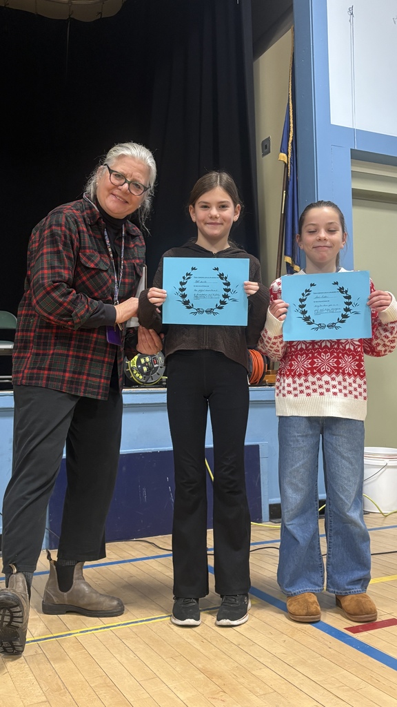 A teacher stands with two elementary students, each holding a blue PBIS classroom award certificate, all smiling on the school stage.