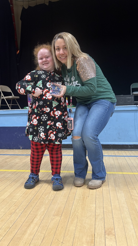 An elementary student smiles while holding a small PBIS specialist award next to a teacher wearing a green holiday-themed shirt during a school assembly.