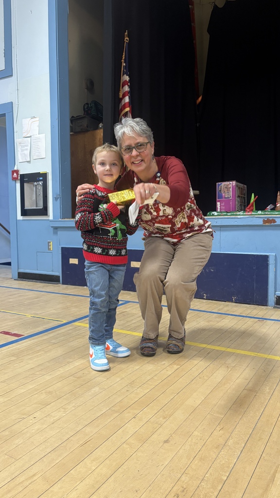 A kindergarten-aged student stands next to a teacher holding a small wrapped award during a PBIS specialist awards presentation on the school stage.