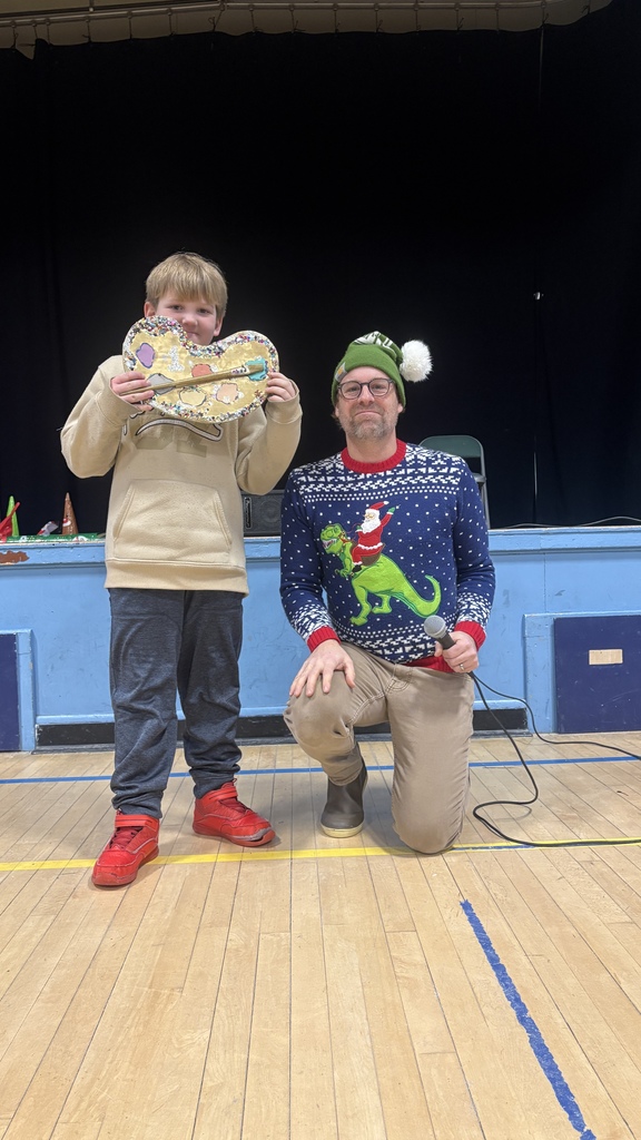 An elementary student holds a decorated guitar-shaped PBIS music award while standing next to a teacher kneeling with a microphone during the school assembly.