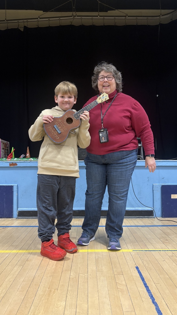 An elementary student stands on the school stage holding a decorated guitar-shaped award while posing with a music teacher, both smiling during a PBIS assembly.