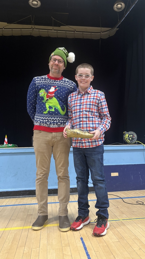 An elementary student wearing glasses holds a gold shoe-shaped PBIS award labeled “MVP” while standing beside a teacher in a festive sweater on the school stage.