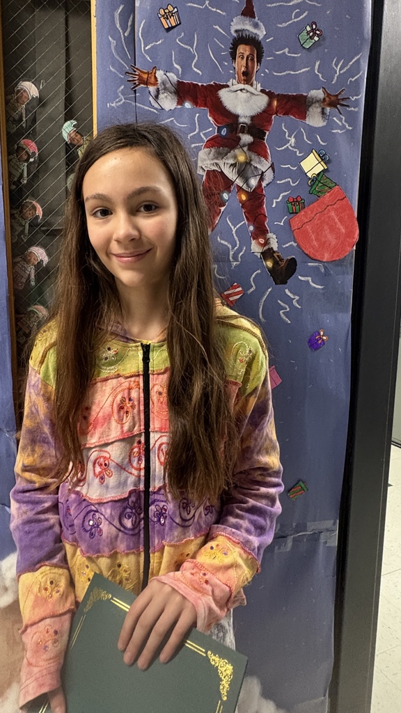 An elementary student stands in a hallway holding a green PBIS award certificate with a festive holiday display in the background.