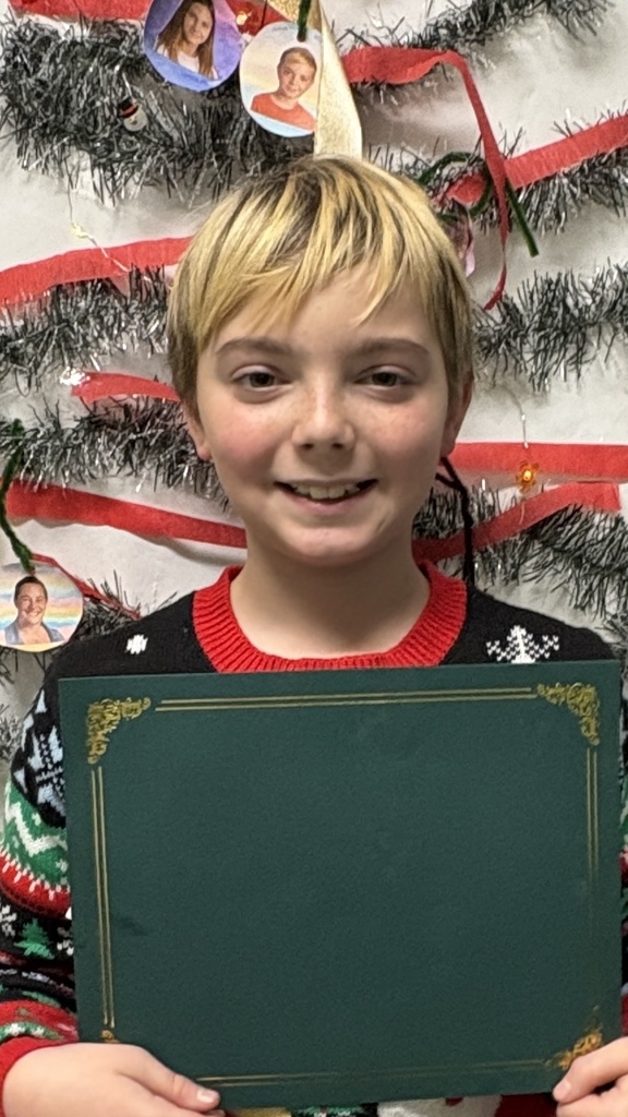 An elementary student holds a green PBIS award certificate in front of a decorated holiday tree display.