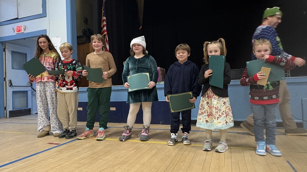 A group of elementary students stand on a school stage holding green PBIS award certificates during an assembly.