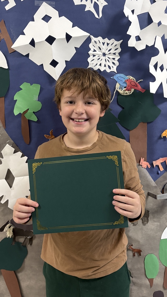 An elementary student smiles while holding a green PBIS award certificate in front of a winter bulletin board with trees and paper snowflakes.