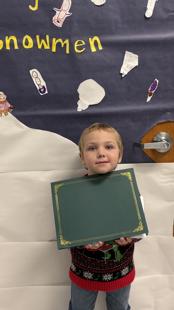 A young elementary student holds a green PBIS award certificate in front of a winter-themed hallway display.