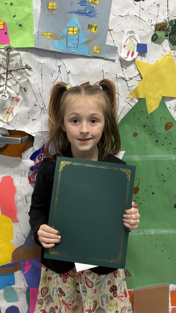 An elementary student with pigtails holds a green PBIS award certificate in front of a colorful classroom art display.