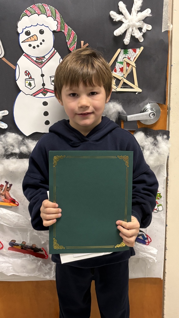 An elementary student holds a green PBIS award certificate in front of a winter-themed classroom display with a snowman.