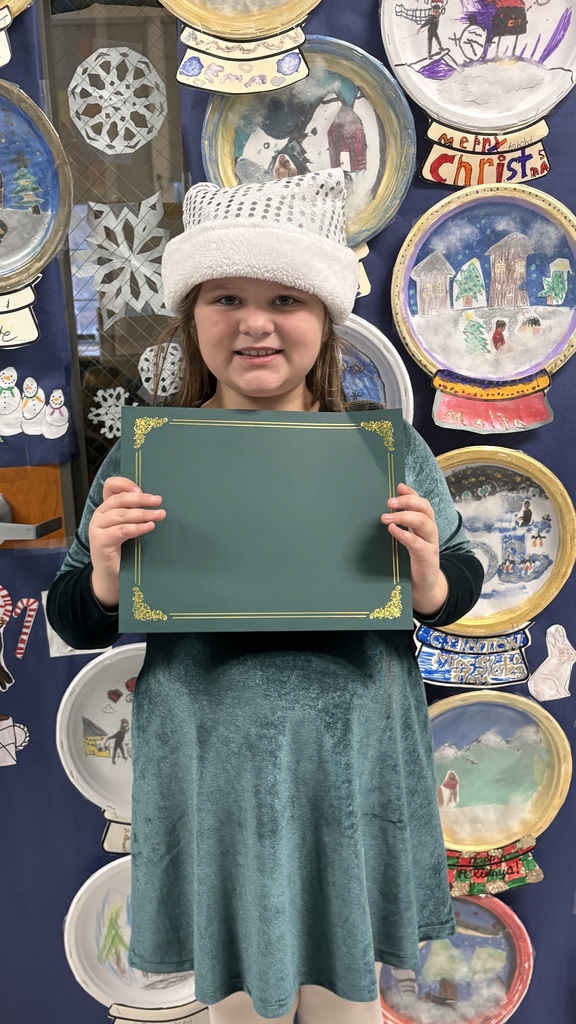 An elementary student wearing a winter hat holds a green PBIS award certificate in front of a display of student snow globe artwork.