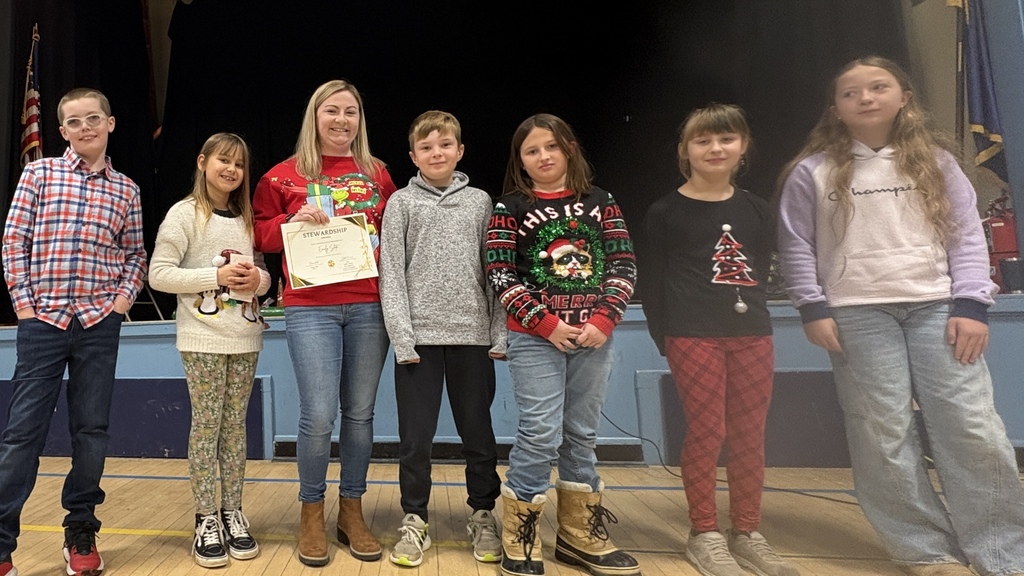 Mrs. Slote stands on a school stage holding a Stewardship Award certificate, surrounded by Student Council members during a school assembly.