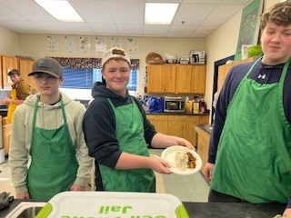 Three students wearing green aprons stand in a kitchen classroom, one holding a plate of food.