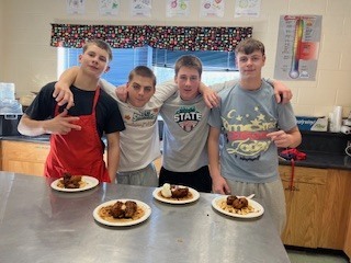 Four students pose together in a kitchen classroom holding plates of food from a cooking competition.