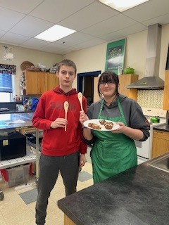 Two students wearing aprons stand in a kitchen classroom holding plates of food during a cooking competition.