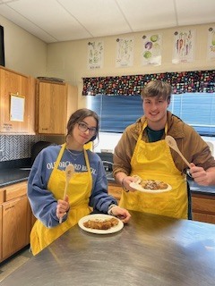Two students wearing aprons stand in a kitchen classroom holding plates of food during a cooking competition.