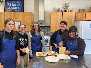 A group of students in aprons stand behind a kitchen counter with finished dishes displayed in front of them.