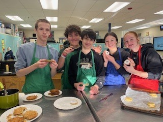 A group of students wearing aprons smile and hold food items while standing around a kitchen worktable.