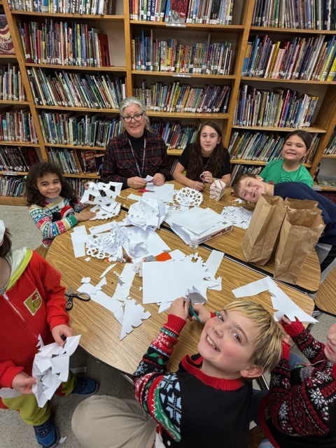 Students and a teacher sit around a table cutting paper snowflakes during a craft activity.