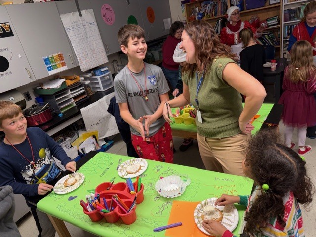 Students and a teacher stand around tables decorating paper plate treats during a holiday classroom activity.