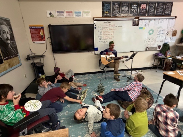 A teacher plays a guitar while students sit and listen on the classroom floor.