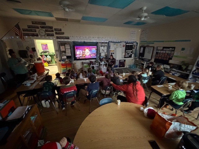 Students sit at tables watching a video on a classroom screen during a holiday activity.