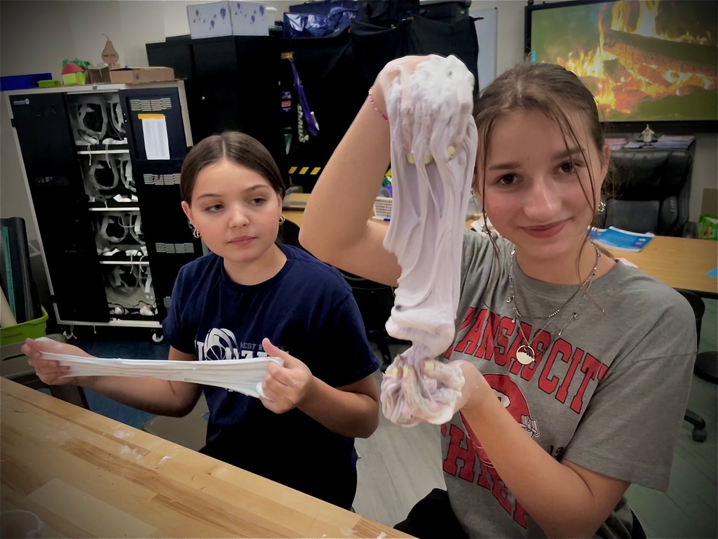 Two students stretch and examine white slime they made at a classroom worktable.