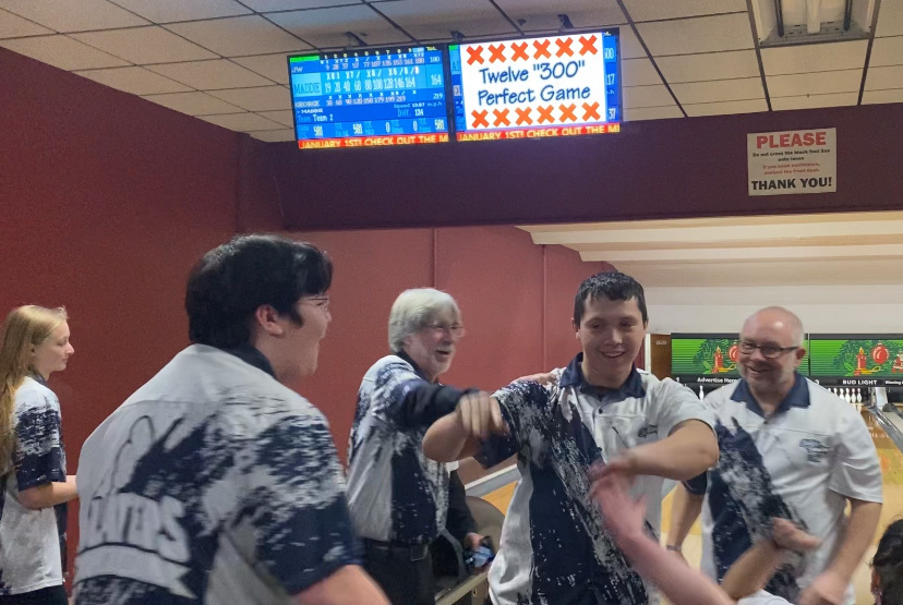 Bowling teammates celebrate with high-fives around a bowler after a perfect 300 game, with the scoreboard overhead reading “Twelve ‘300’ Perfect Game” at Rutland Bowlerama.