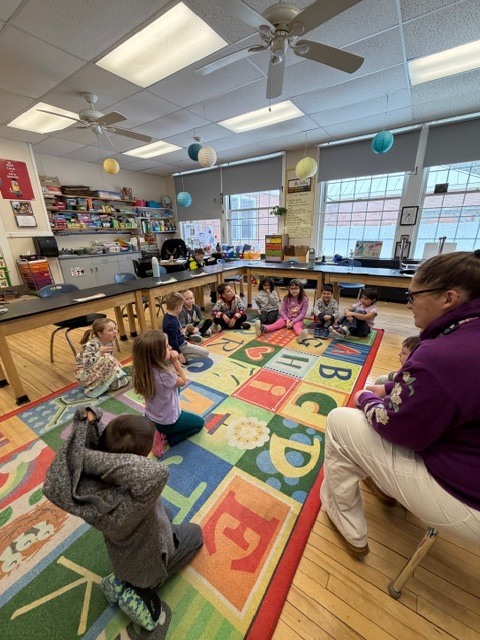 Kindergarten story time: Young students sit on a colorful alphabet rug while a teacher reads aloud to the class.