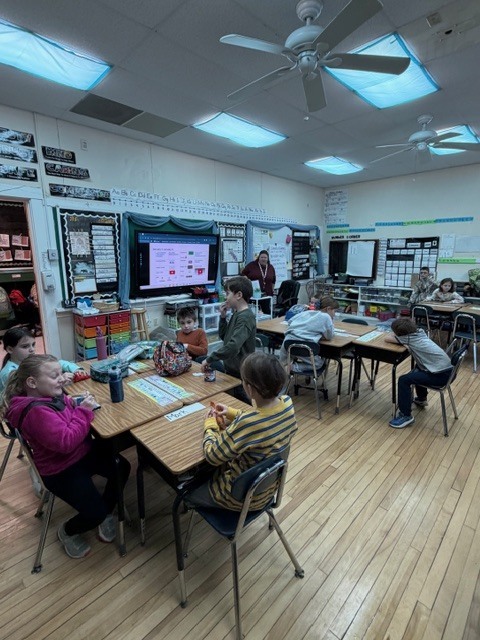Students working at desks: Elementary students sit at desks completing assignments while a teacher provides instruction at the front of the room.
